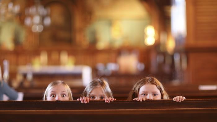 Tre flickor tittar busigt upp bakom en kyrkbänk.