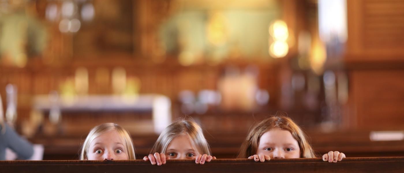 Tre flickor tittar busigt upp bakom en kyrkbänk.
