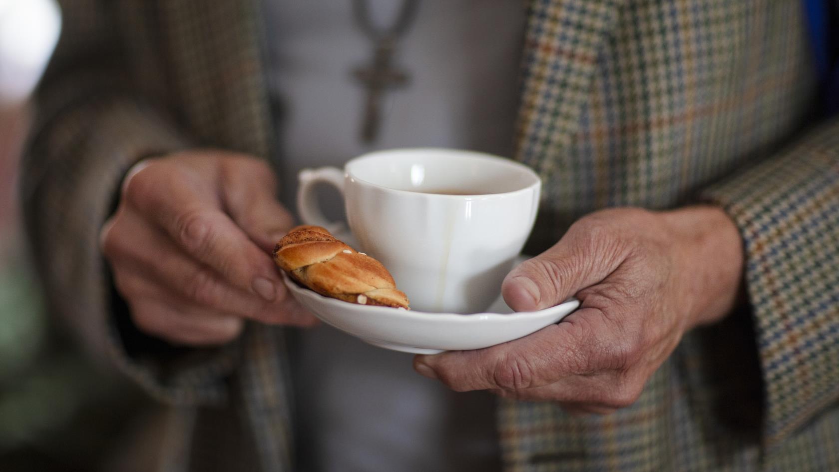 Någon i rutig kavaj håller en kaffekopp och en kanelbulle på fatet.