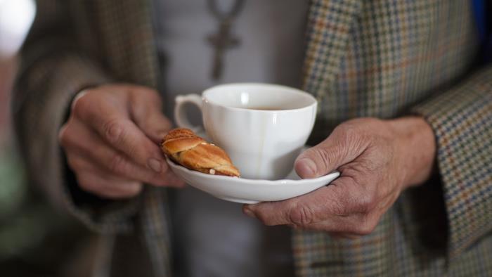 Någon i rutig kavaj håller en kaffekopp och en kanelbulle på fatet.