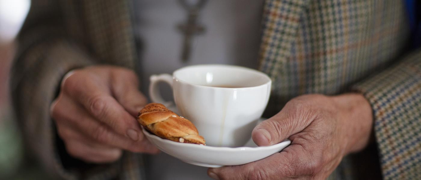 Någon i rutig kavaj håller en kaffekopp och en kanelbulle på fatet.