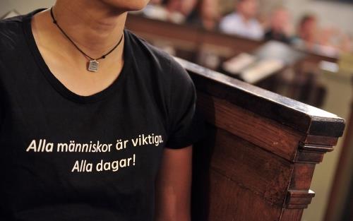 En person i svart t-shirt med texten Alla människor är viktiga alla dagar, sitter i en kyrkbänk.