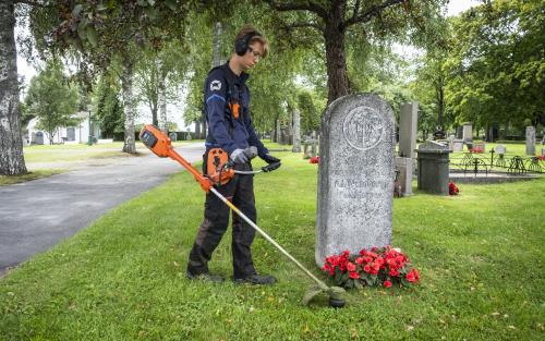 En ung kille klipper gräset runt gravstenarna med en trimmer på en kyrkogård.