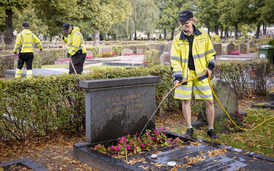 En kyrkogårdsarbetare i gula varselkläder vattnar blommorna på en gravplats.