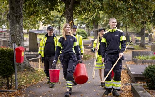 Några kyrkogårdsarbetare i gula varselkläder har olika trädgårdsredskap i händerna.