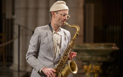 En man i ljusgrå kostym och vit mössa spelar saxofon i Uppsala domkyrka.
Foc