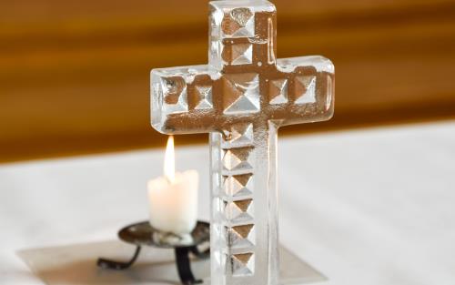 Närbild på ett litet glaskors och ett tänt ljus på ett altare.