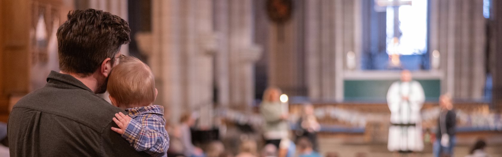 Pappa står längst bak i kyrkan med ett litet barn i famnen. Han tittar mot prästen som pratar inför en fullsatt kyrka.