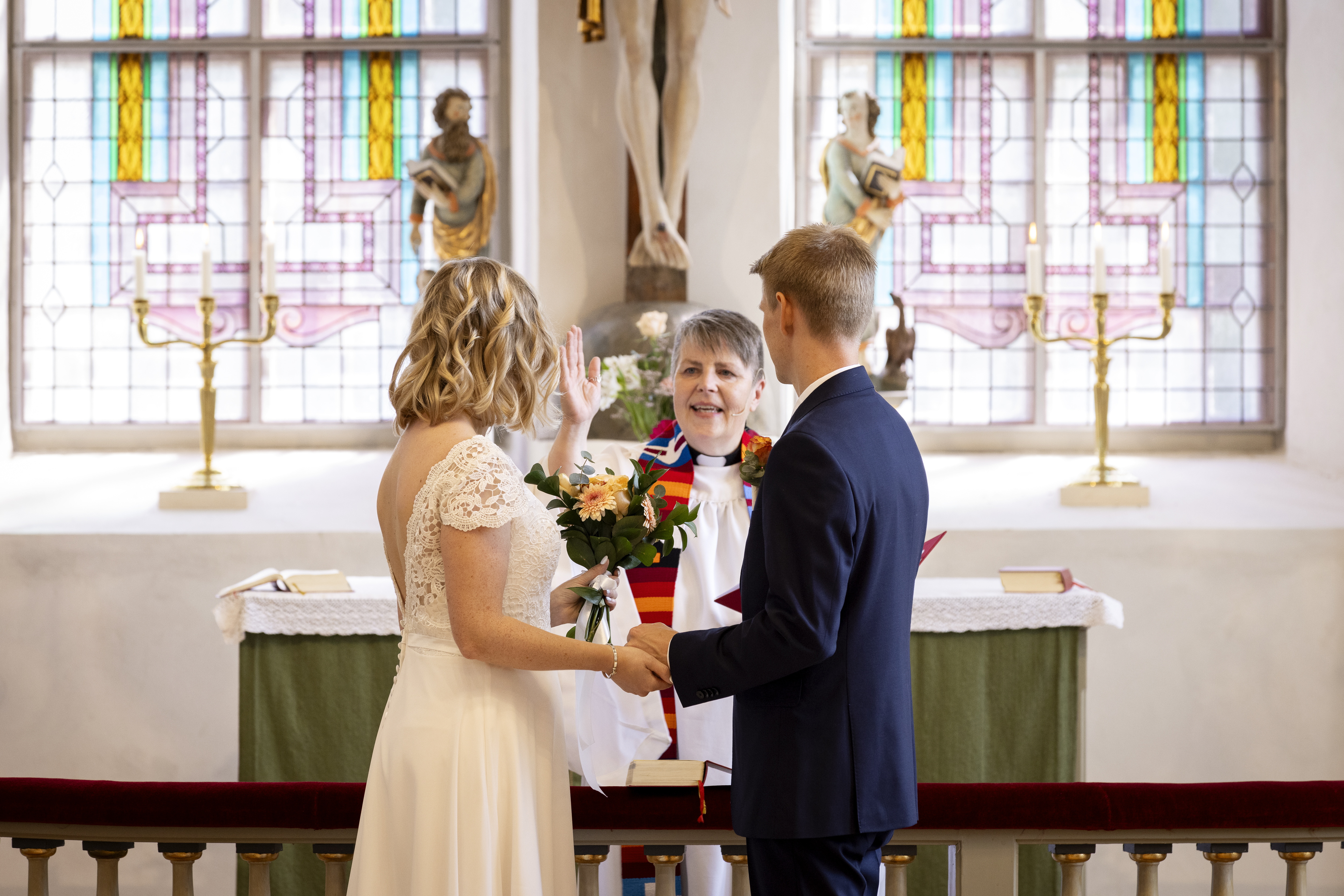 Drop-in vigsel i en kyrka. Ett brudpar står framför en präst vid altarringen. Bruden håller en blombukett i handen.