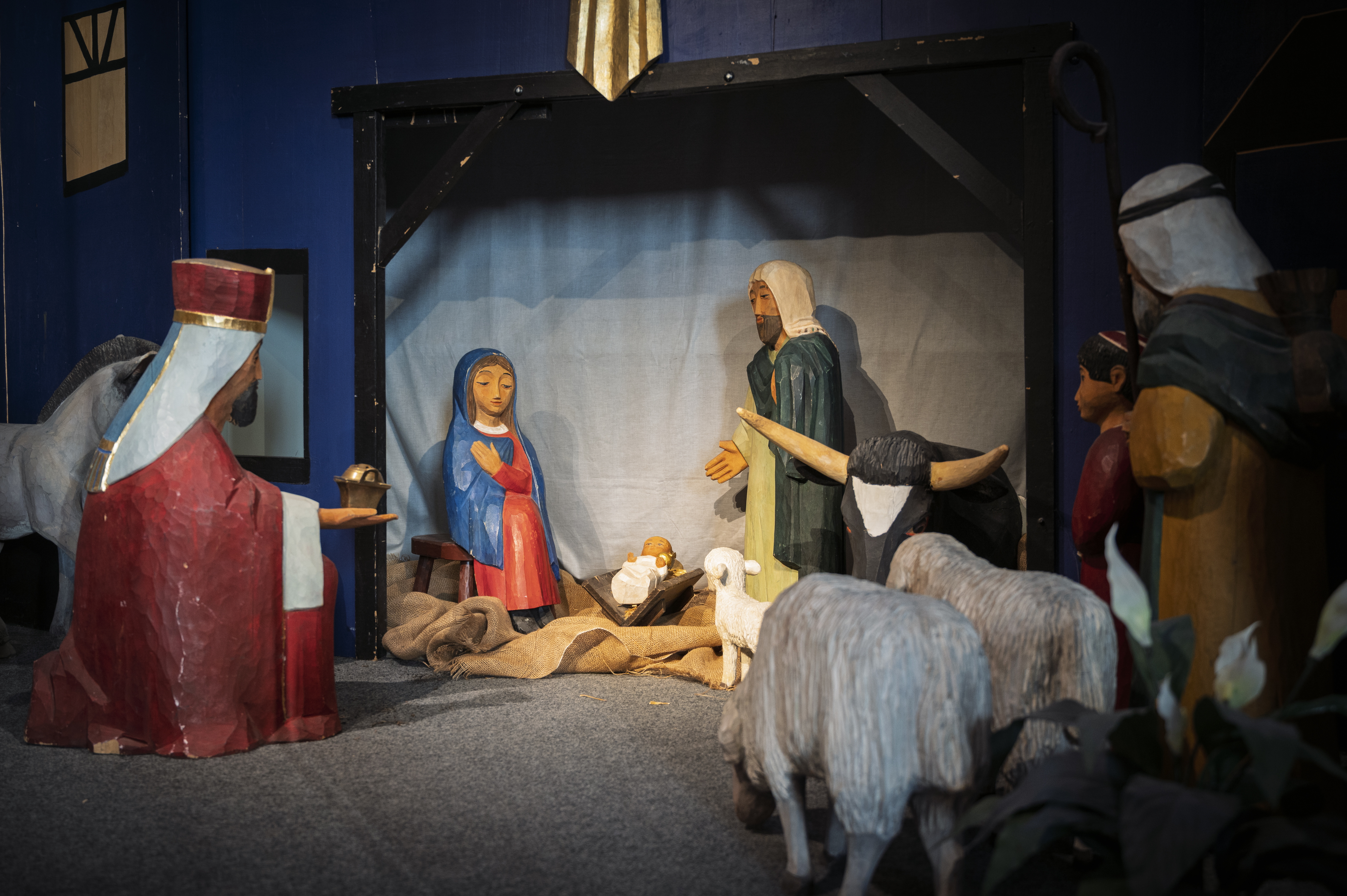 En upplyst julkrubba. Trädockor som föreställer Josef, Maria och Jesusbarnet i krubban. De tre vise männen och får i förgrunden.