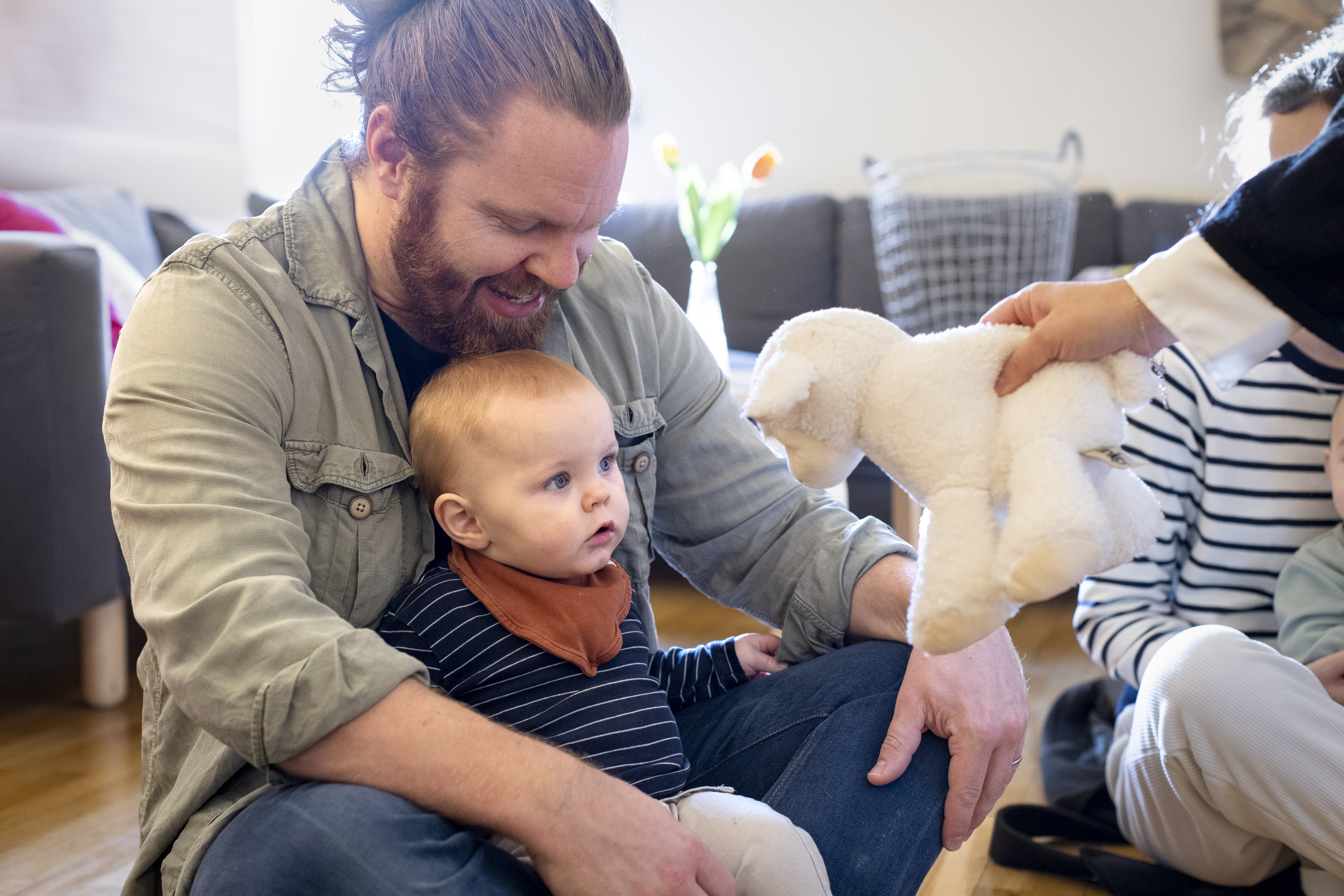 En man i skägg sitter på golvet i skräddarställning med ett litet barn i knät. En person sträcker fram ett gosedjurs-lamm mot barnet.