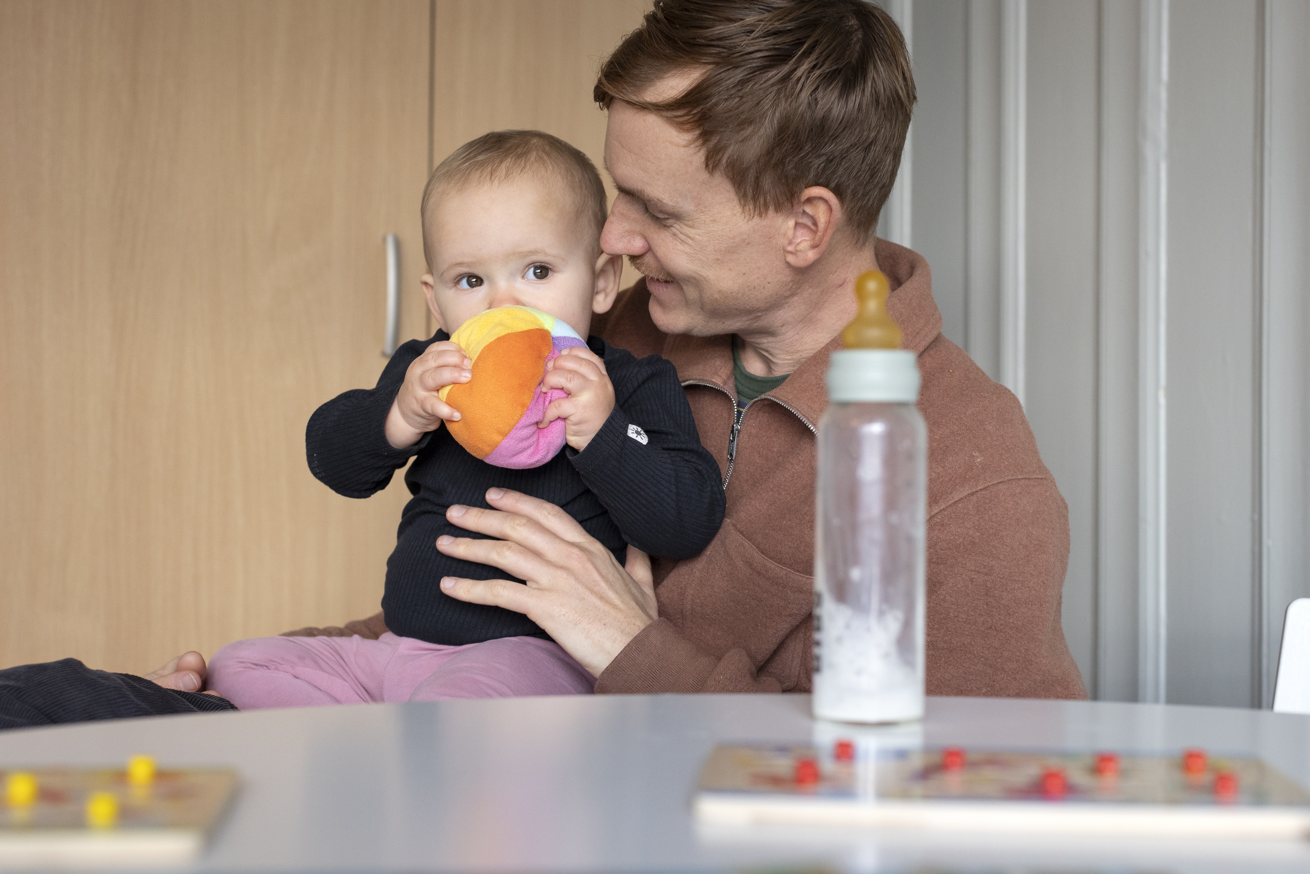 En man sitter med ett litet barn i knät. Barnet håller en mjuk färgglad boll med båda händerna framför munnen.
