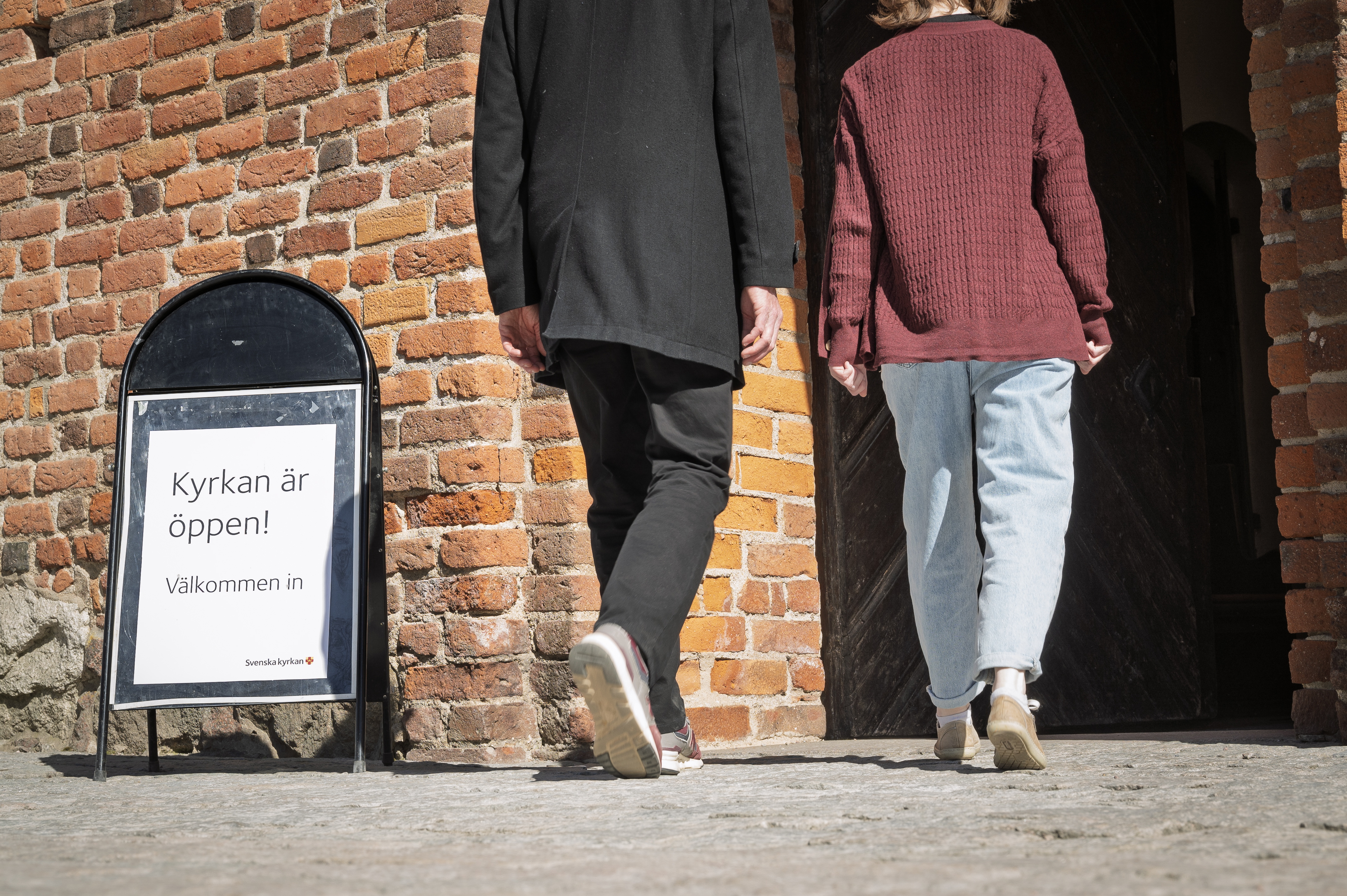Två personer på väg in i en kyrka. En skylt med texten att kyrkan är öppen står bredvid dörren.