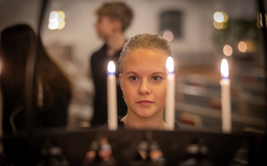 En tjej står framför en ljusbärare med tända ljus. I bakgrunden står några ungdomar.