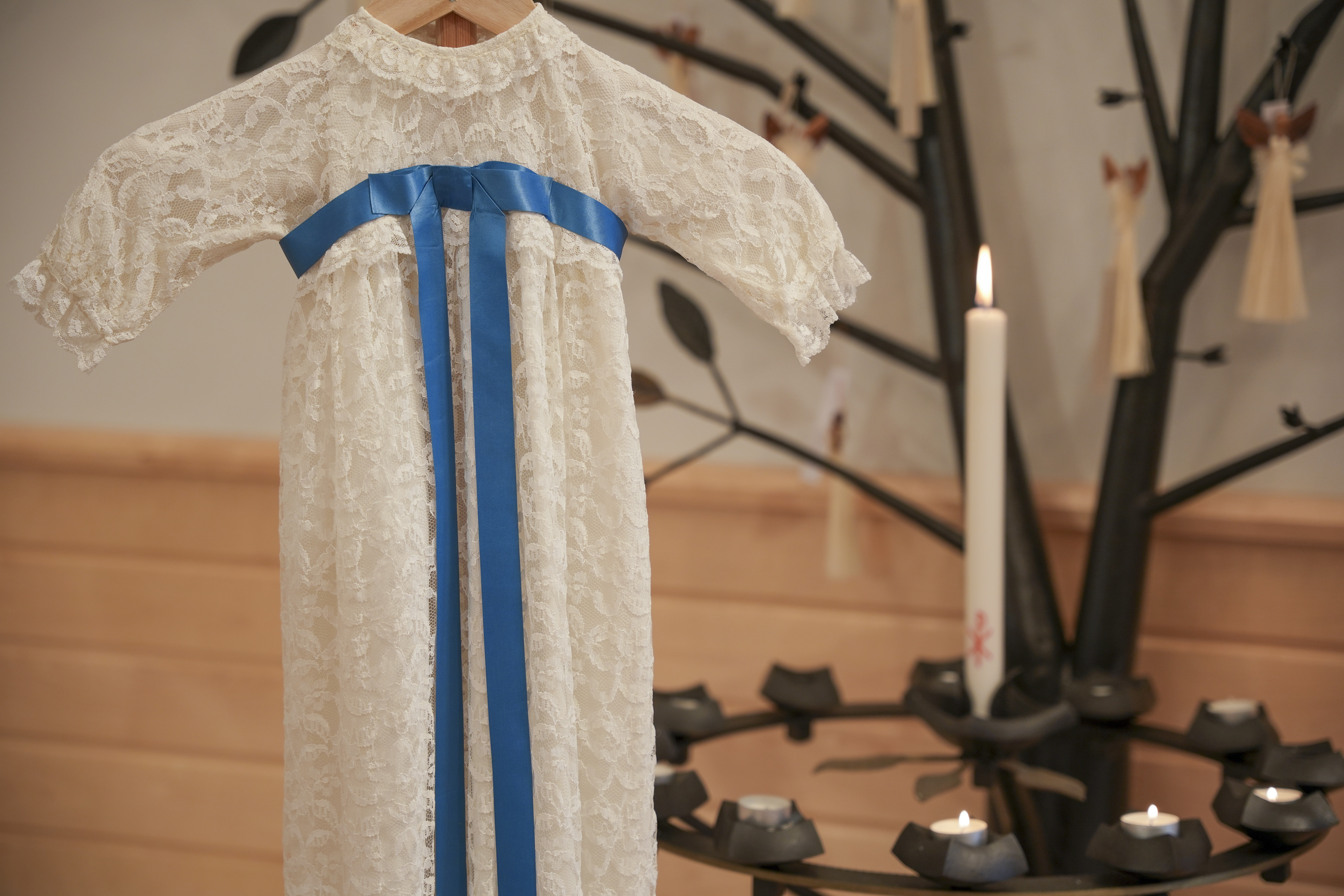 En dopklänning i vit spets med blått band. Ett dopljus brinner i bakgrunden.