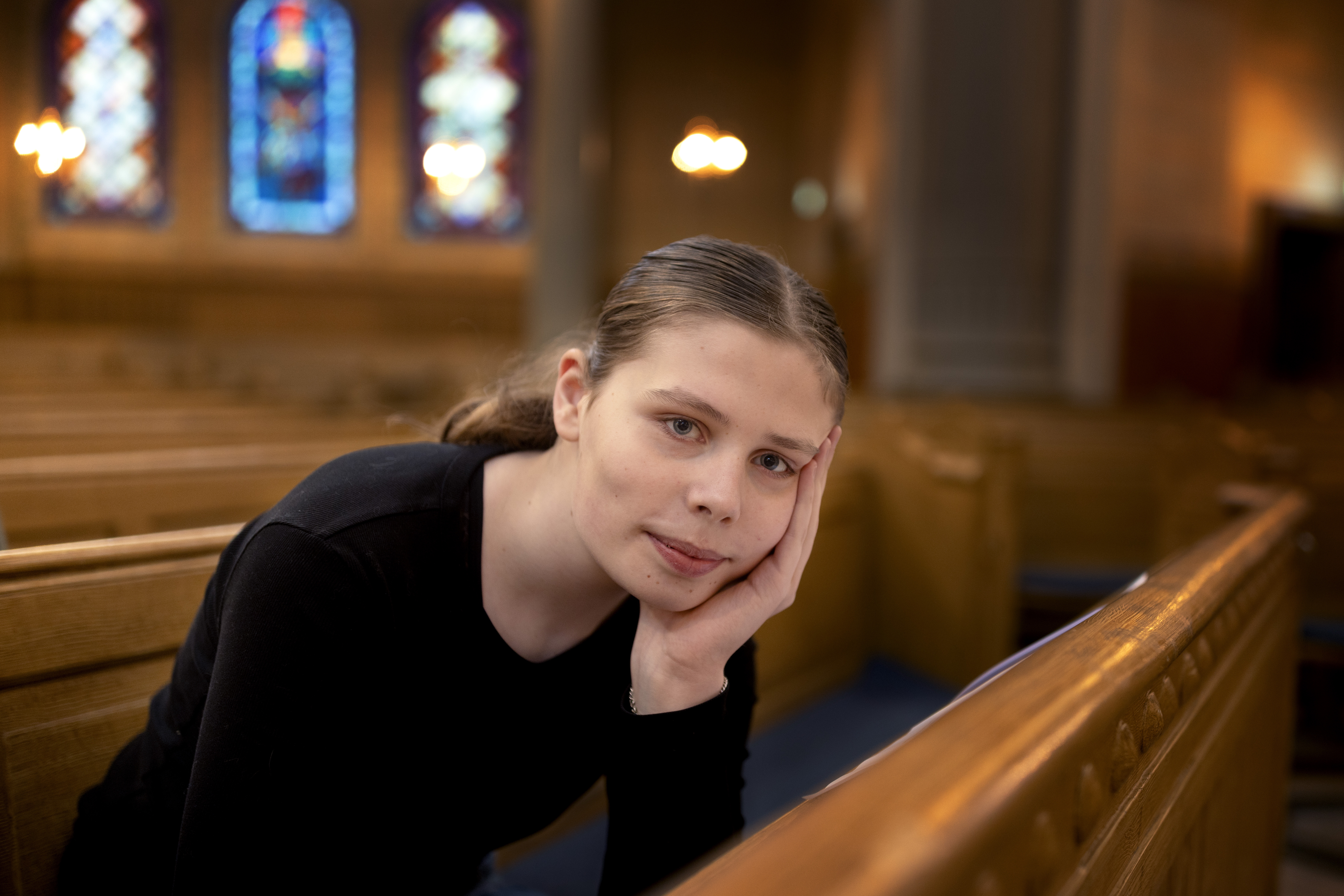 Närbild på en yngre kvinna som sitter i en kyrkbänk och lutar sin kind mot handen.