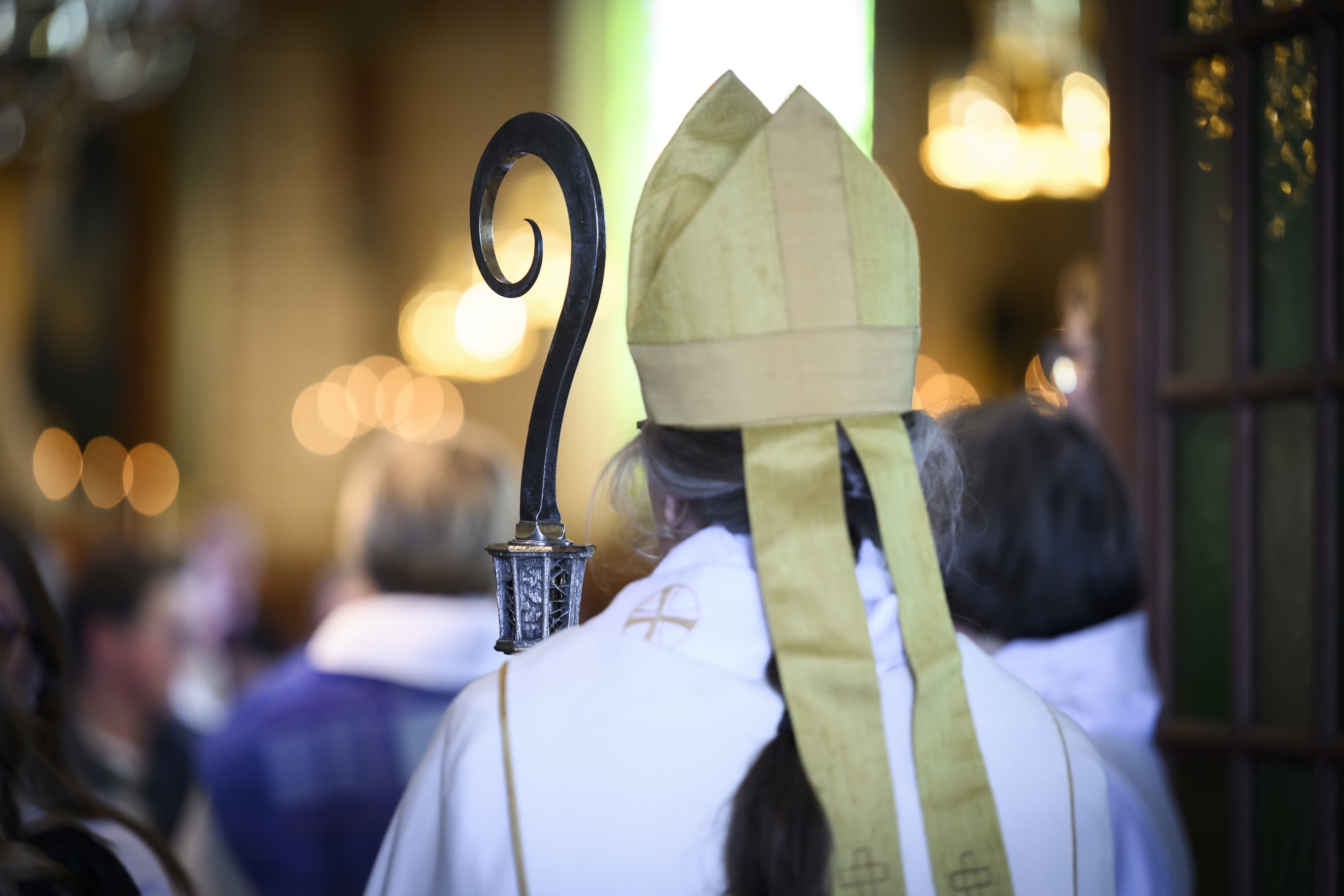 En biskop med ryggen mot kameran på väg in i en kyrka. Hen bär en guldfärgad mitra och håller en kräkla i handen.