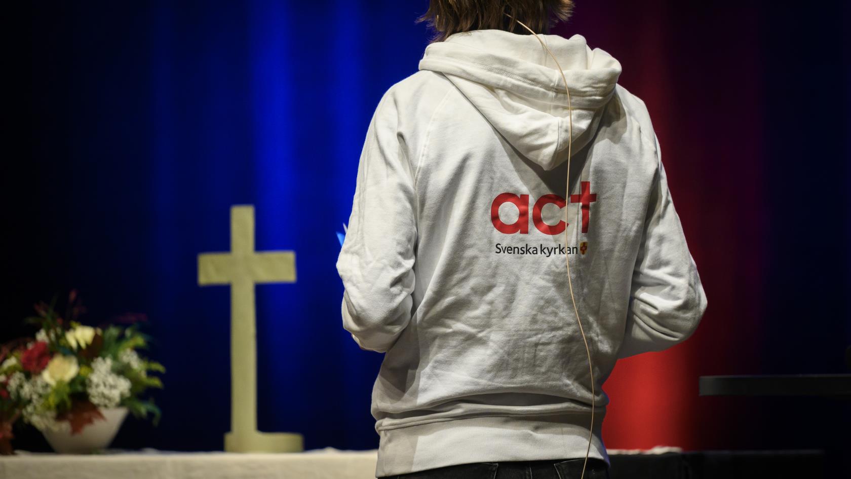 Altare med ett kors. Tyggtavlan av en person i luvtröva med texten Act Svenska kyrkan på ryggen. 