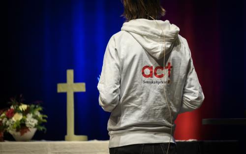 Altare med ett kors. Tyggtavlan av en person i luvtröva med texten Act Svenska kyrkan på ryggen. 