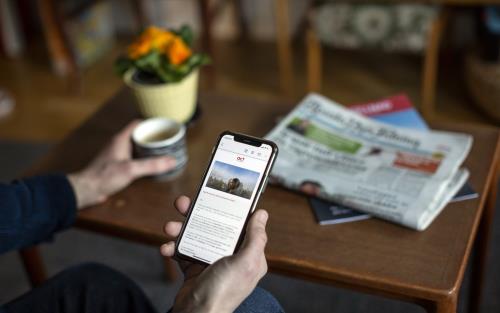 Ser en hand som håller i en mobiltelefon och läser ett nyhetsbrev. På soffbordet bakom står en kaffekopp och en liten hög med papperstidningar.