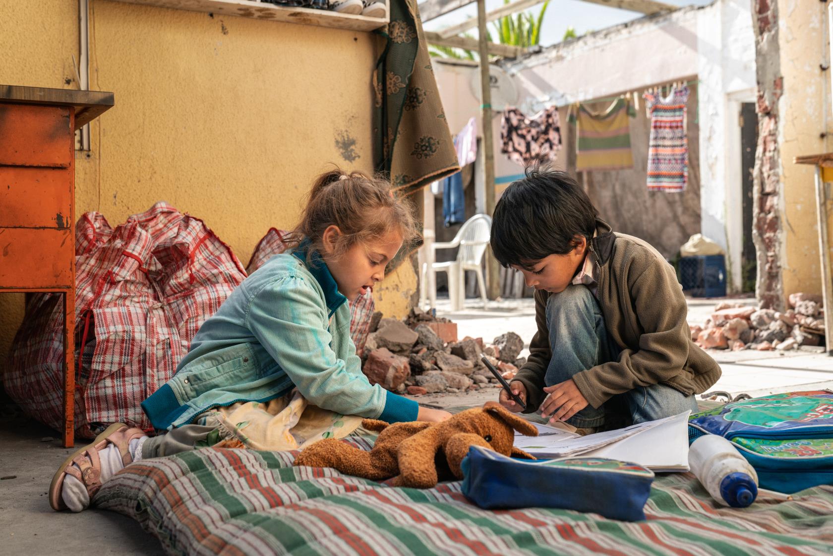 Två barn sitter på golvet på en madrass och skriver. Framför dem ligger skolböcker och en nalle.