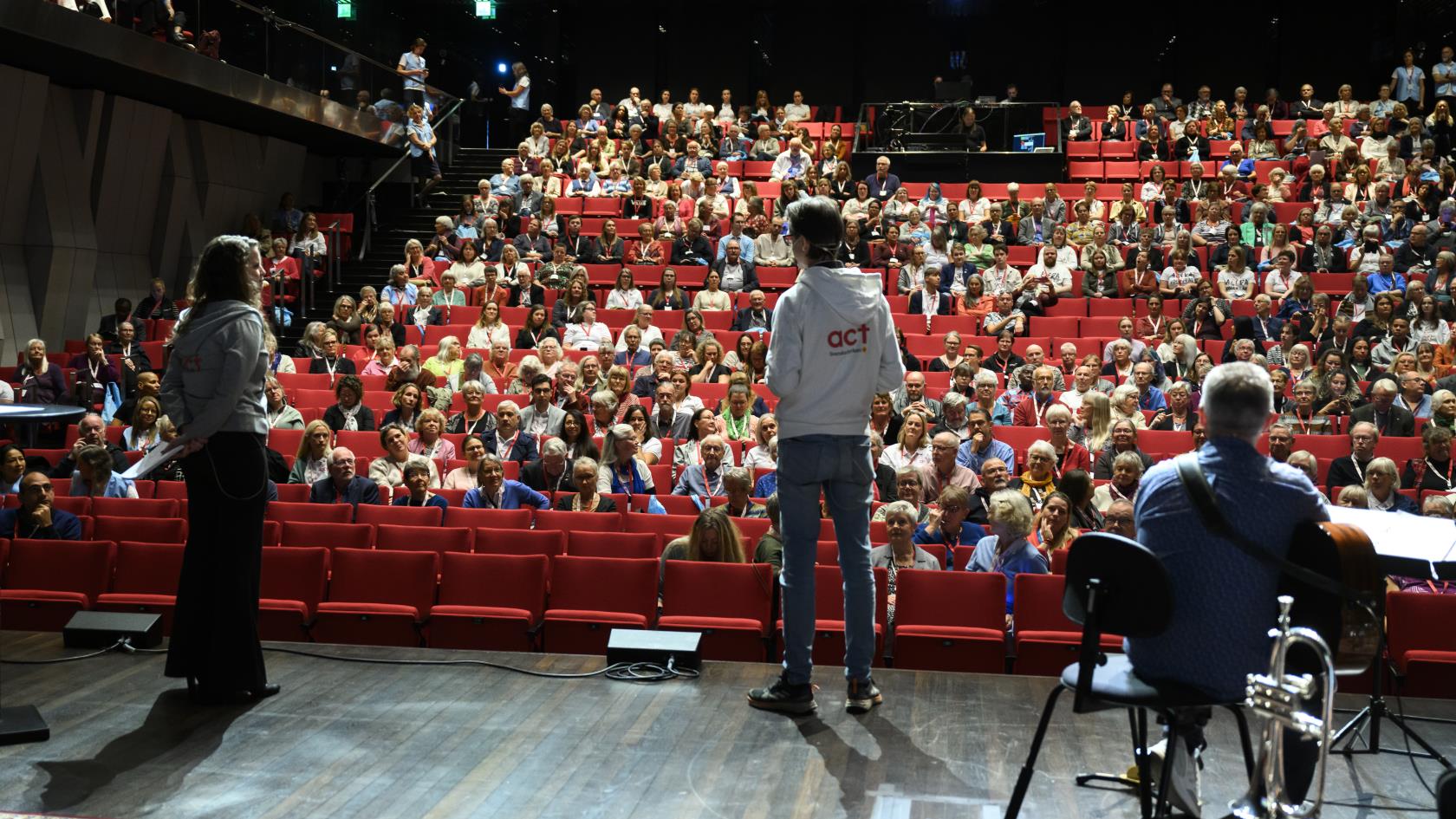 Ryggar på tre personer på en scen. I publiken ser vi flera hundra åskådare. 