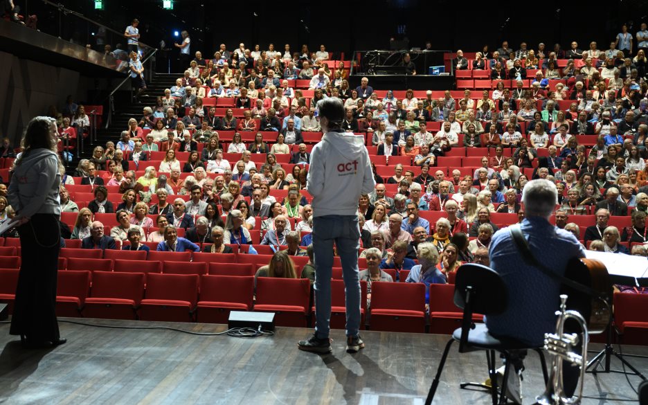 Ryggar på tre personer på en scen. I publiken ser vi flera hundra åskådare.