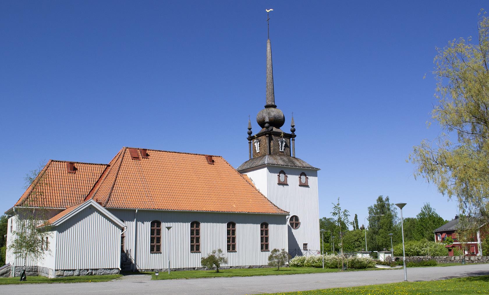 Bilden visar Kåge kyrka i sommarskrud. Kyrkan är målad i ljus grå kulör och har ett rött tegeltak. 