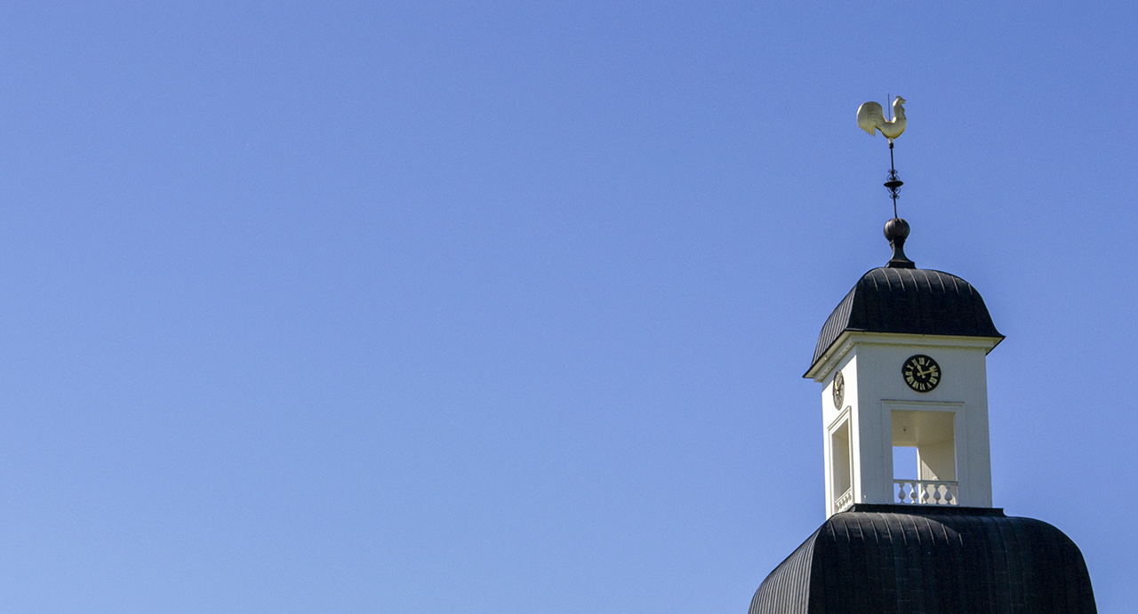 Bilden visar Kusmarks kyrkas torn mot blå himmel. Längs upp i tornspiran sitter den förgyllda kyrktuppen. 