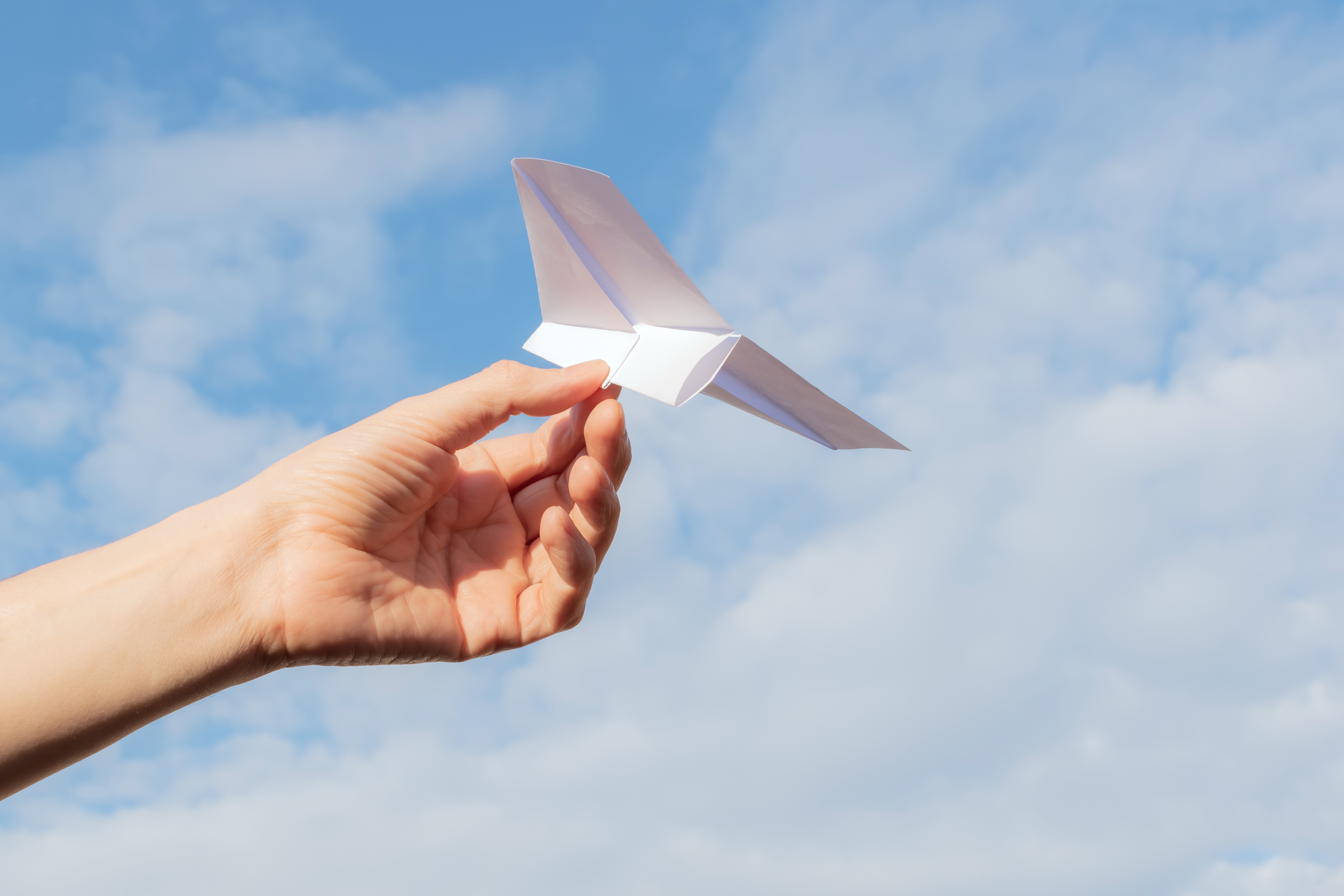 En hand håller ett pappersflygplan och siktar det mot en blå himmel.