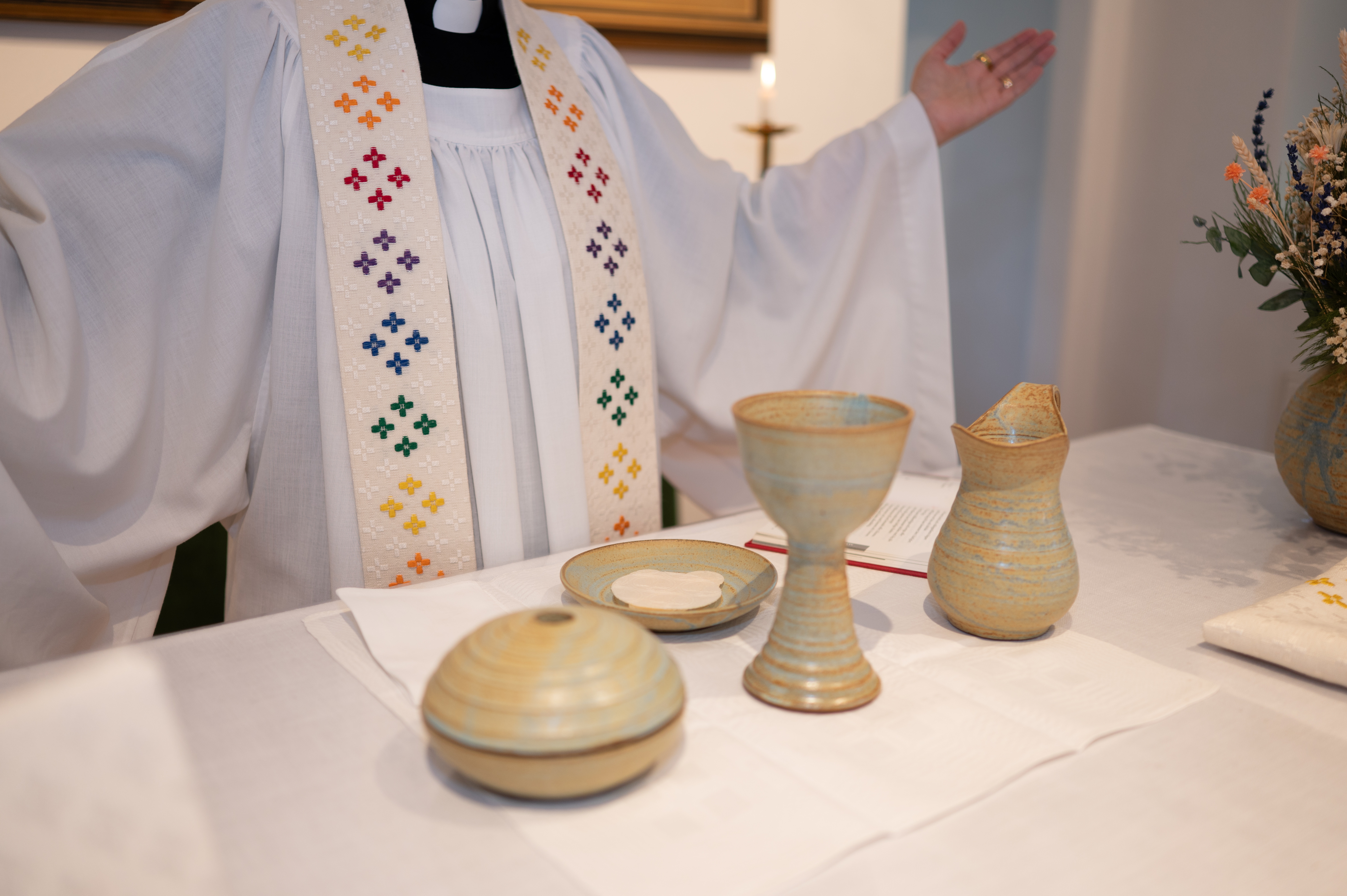 Närbild på keramisk nattvardsservis med vin och oblater. Föremålen står på ett altare, och en präst med röklin och stola skymtar i bakgrunden.