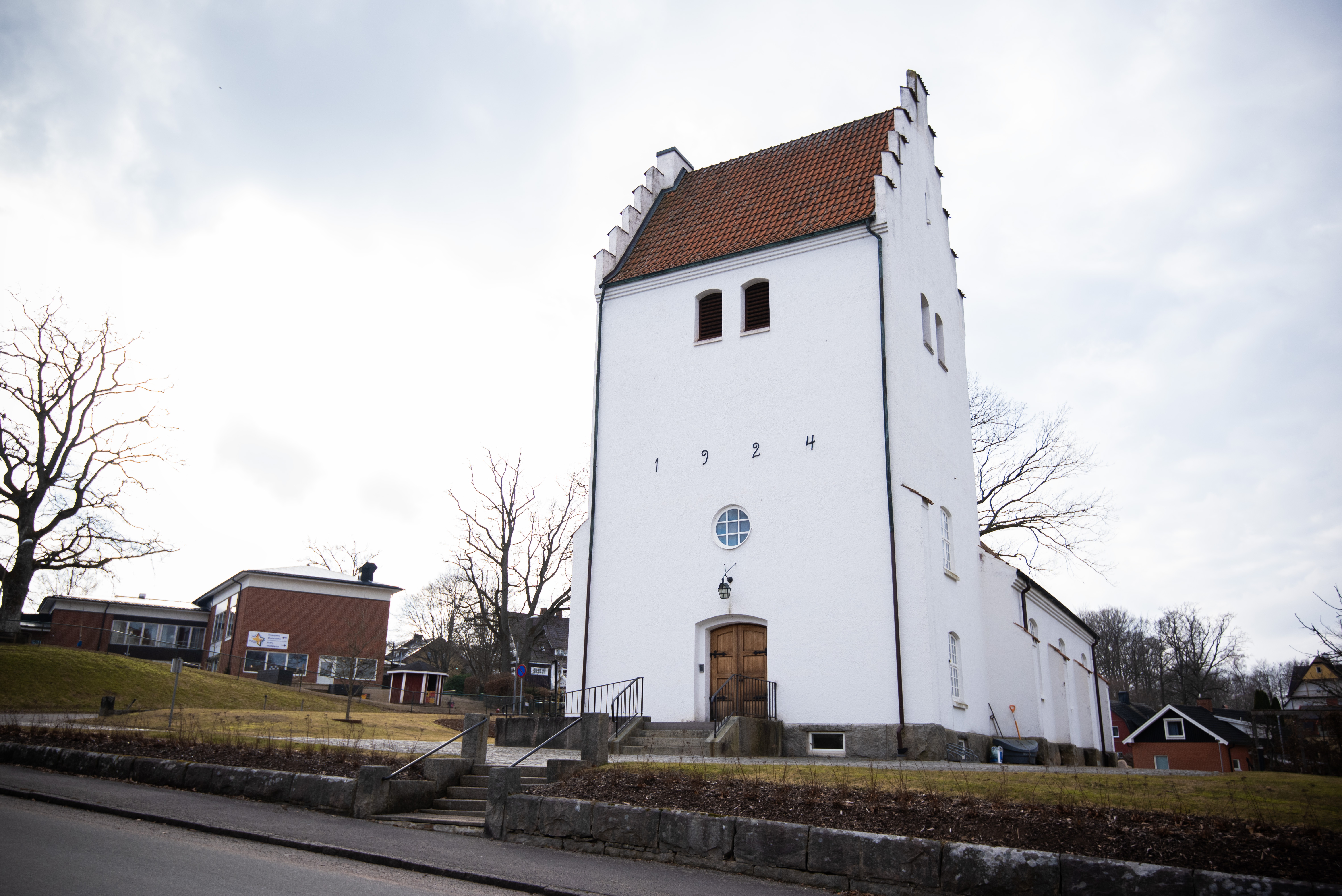 En vit kyrka utmed en gata i en by. I bakgrunden skymtar ett församlingshem med en lekplats.
