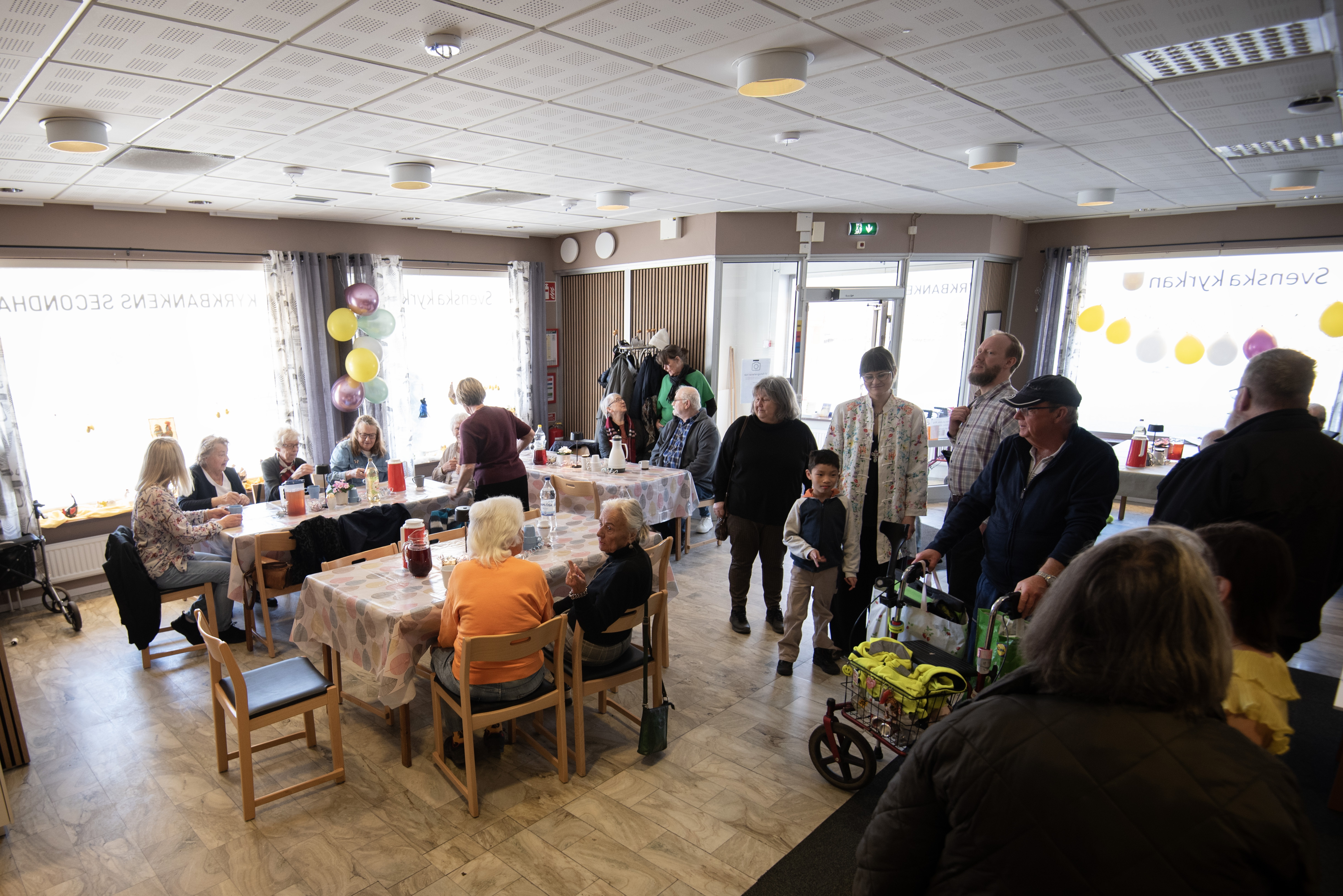 En lokal med öppen golvyta och stora skyltfönster. Lokalen är full av folk. Några sitter vid bord och äter tårta. Det hänger ballonger på väggarna.