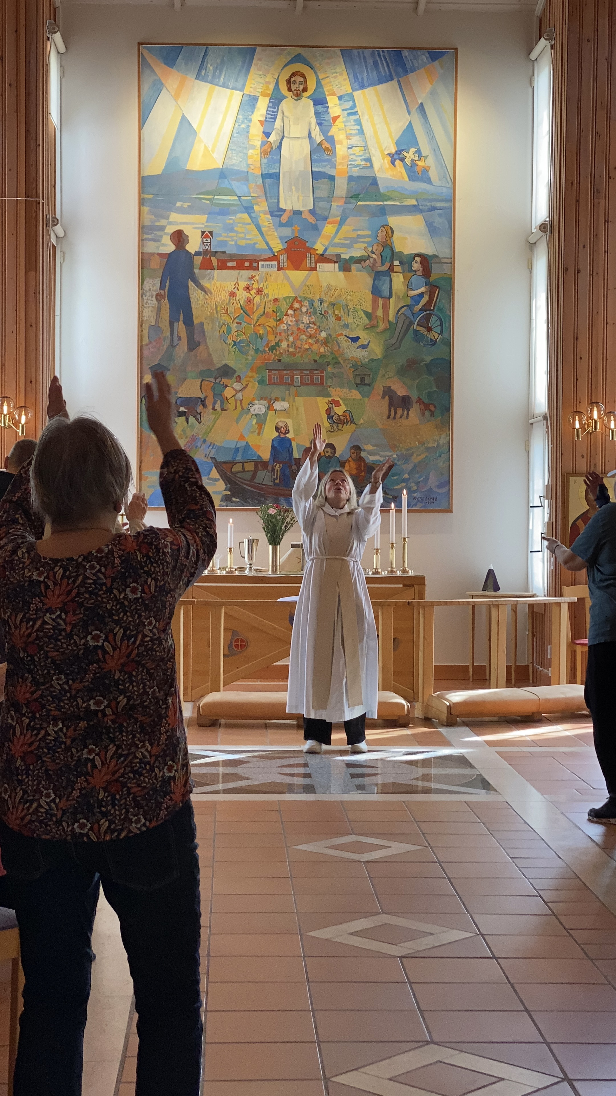 Präst står framför altaret och lyfter sina händer upp över huvudet. I förgrunden ses även gudstjänstdeltagarna göra detsamma. 