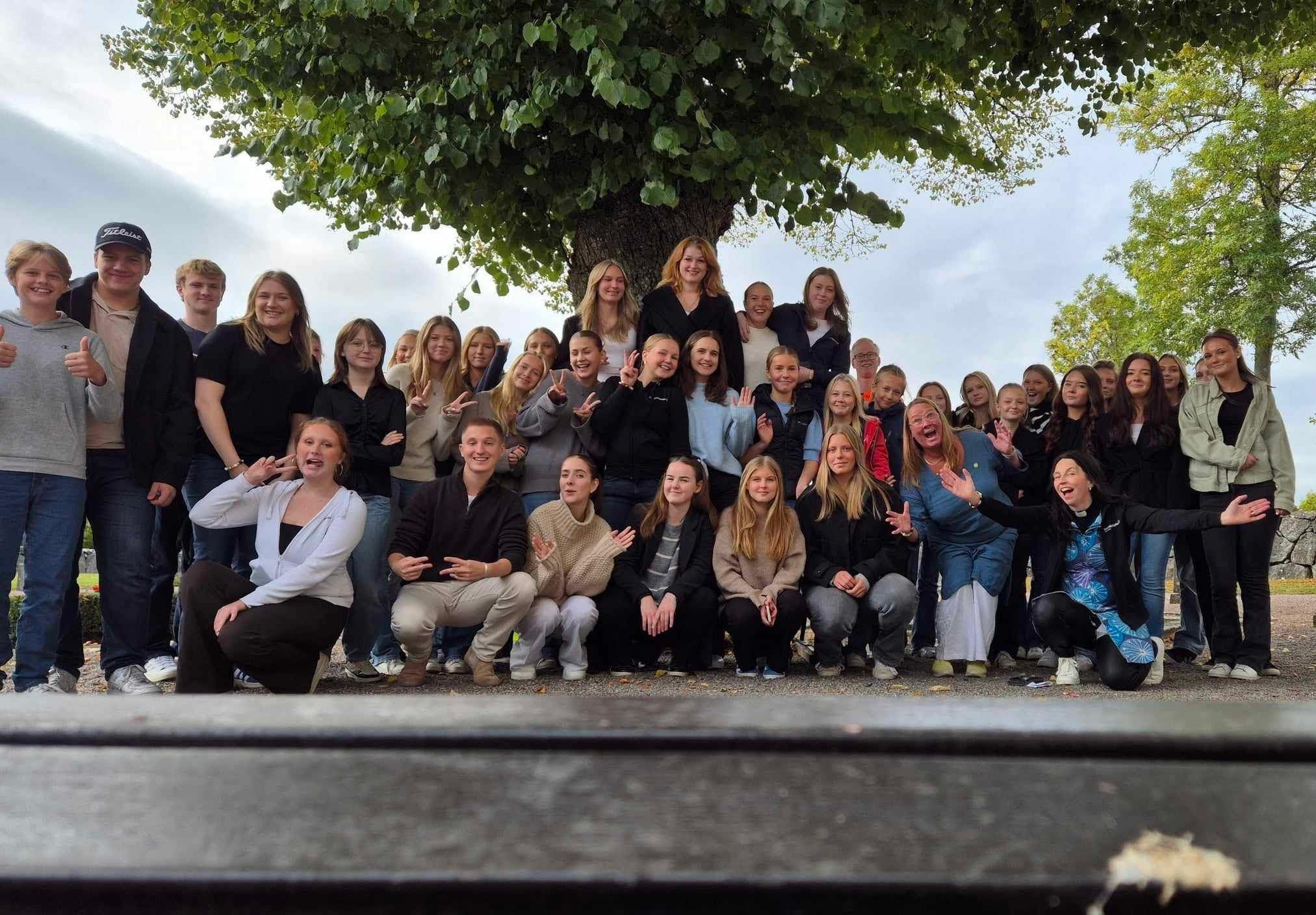 En grupp med runt tjugo tonåringar står uppställda framför kameran. Vissa håller armarna om varandra, vissa gör V-tecken med fingrarna eller räcker ut tungan.