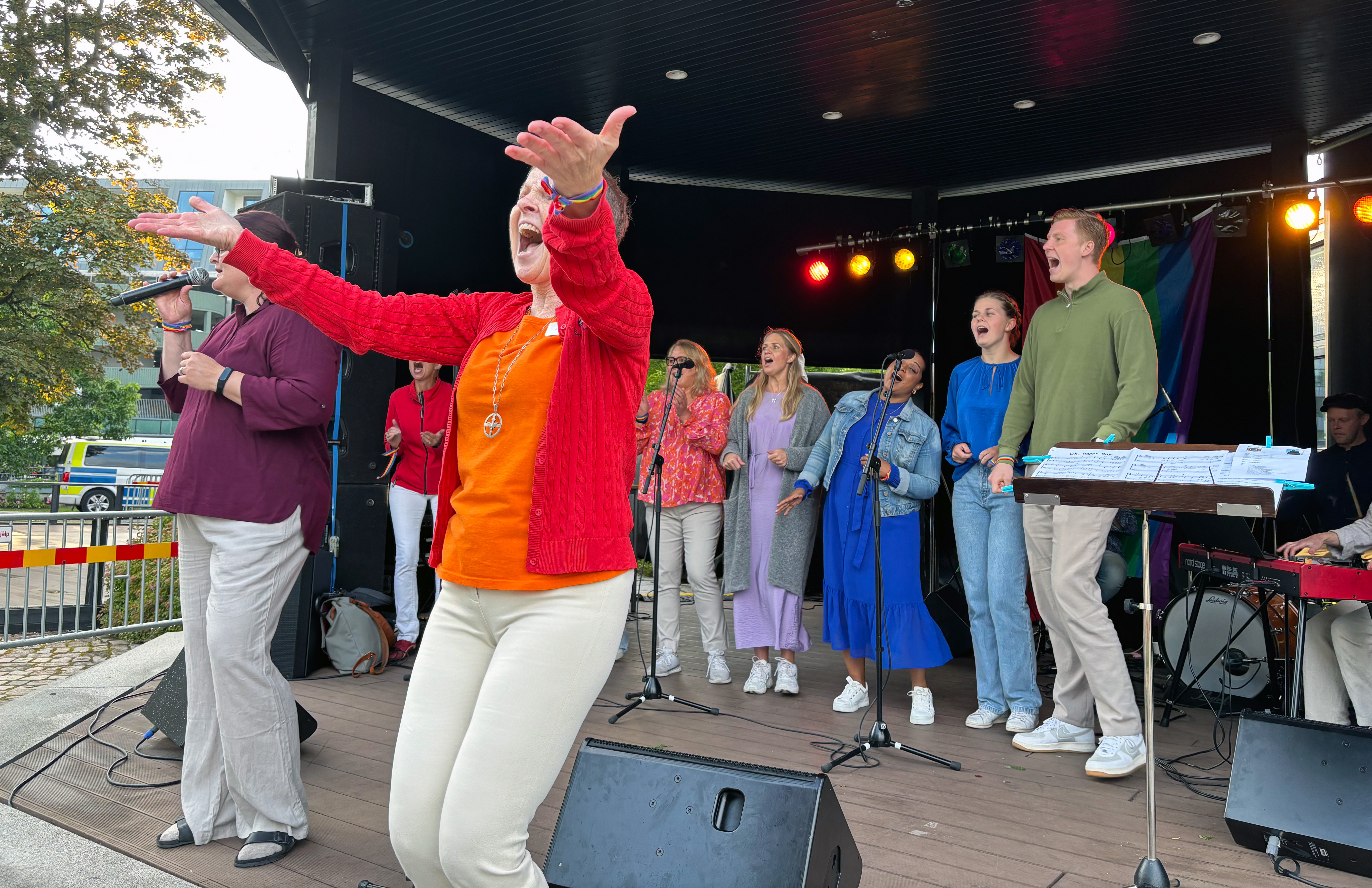 Kvinna i orange och rött leder kör på scen utomhus.