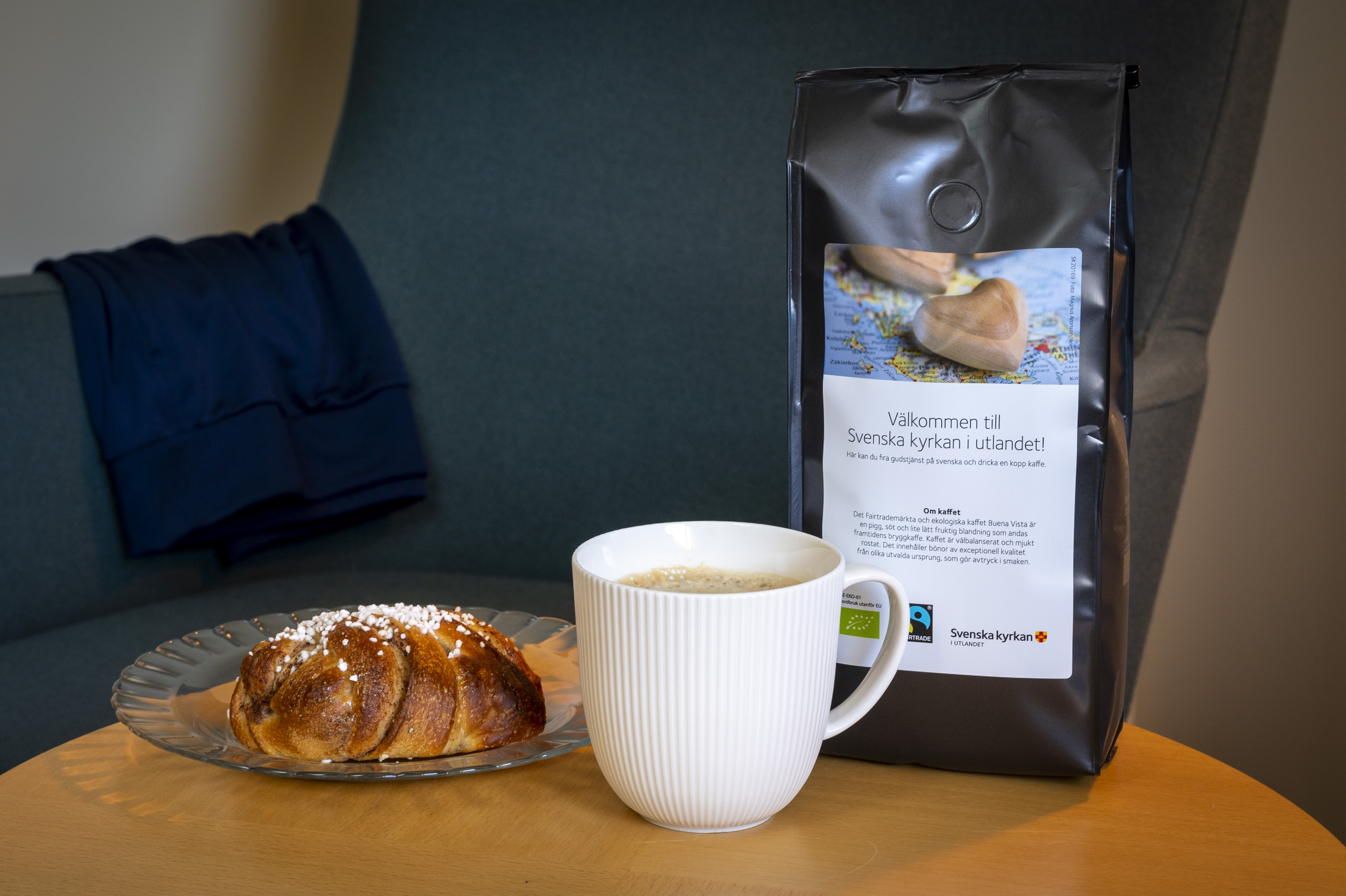 En kaffekopp, en kanelbulle och en påse med kaffe från Svenska kyrkan står på ett bord.