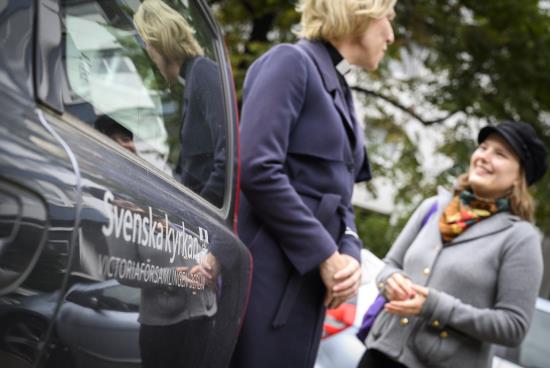 En kvinnlig präst samtalar med en kvinna utanför en bil med Svenska kyrkans logga.
