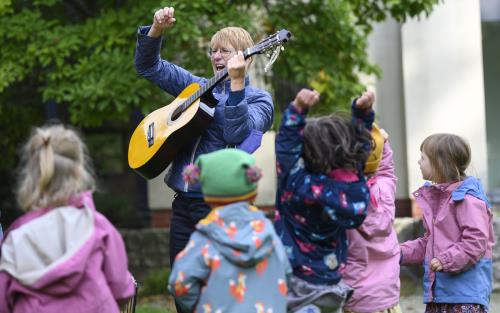 En kvinna står utomhus och spelar gitarr för en grupp barn.