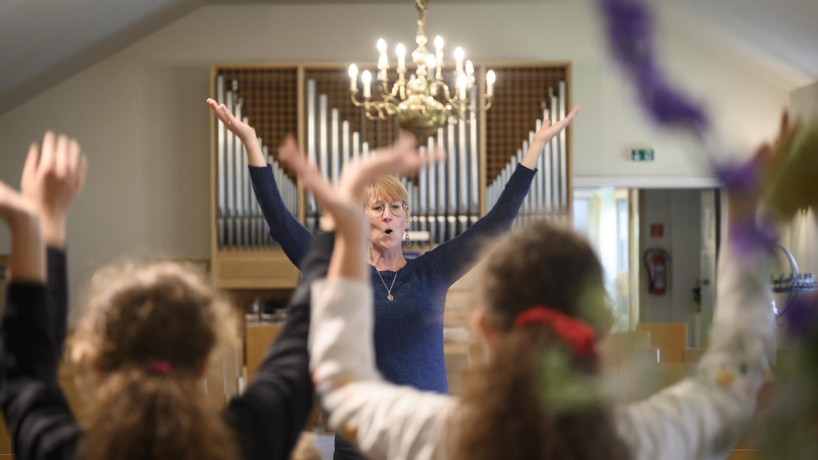 En kvinna står framför en orgel med armarna upp i luften. Några barn står i förgrunden och gör samma gest.