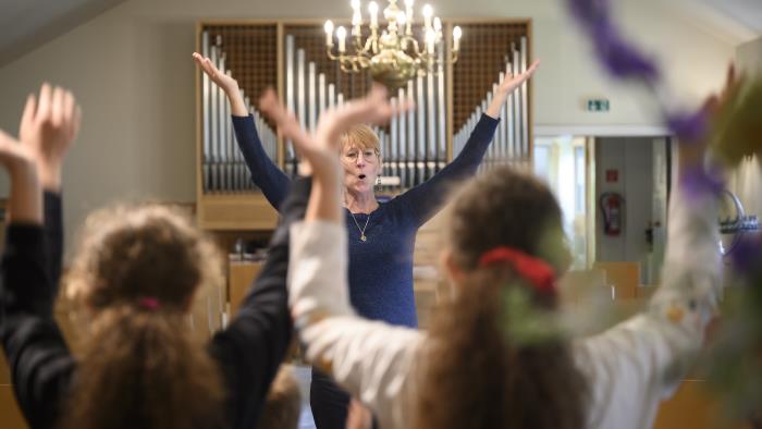 En kvinna står framför en orgel med armarna upp i luften. Några barn står i förgrunden och gör samma gest.