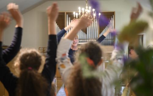Några ungdomar står i en kyrka och viftar med armarna i luften. En kvinna står framför dem och gör likadant.