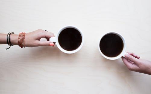Två händer håller i varsin kopp med kaffe som står på ett bord.