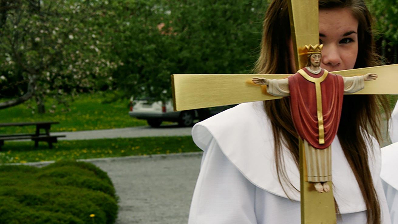 Konfirmand med processionskors med Kristusfigur på. Hennes ansikte är lite dolt bakom korset.