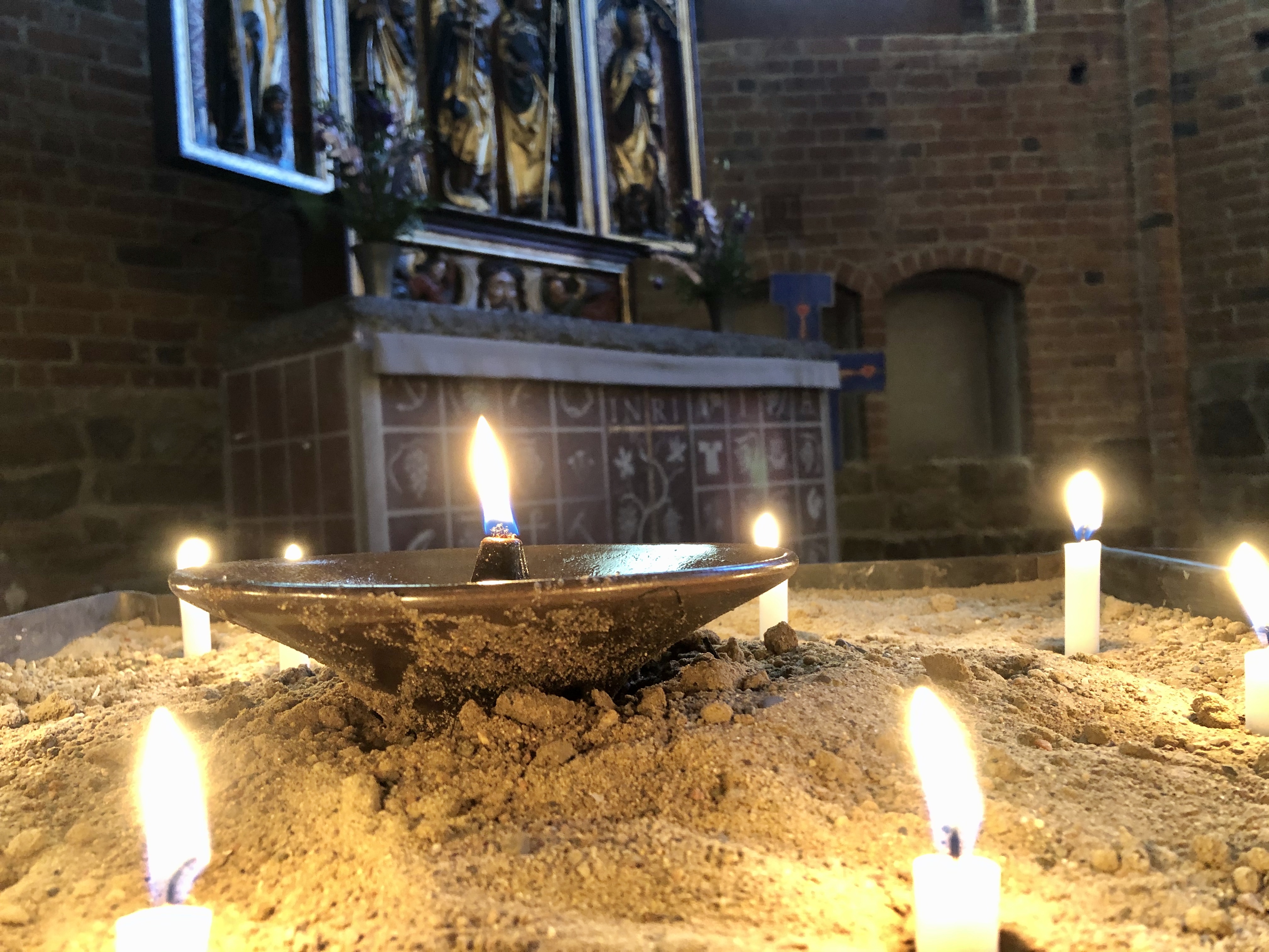 Oljelampa som står i en sandljusbärare. Runt oljelampan är små stearinljus tända i sanden. I bakgrunden skymtas Klosterkyrkans högaltare och altartavla.