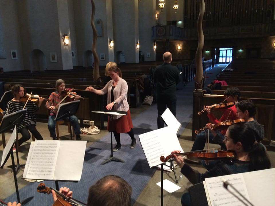 Ett antal fiolspelande personer sitter med notblad framför sig i en cirkel i kyrkan och en dirigent leder musiken. 
