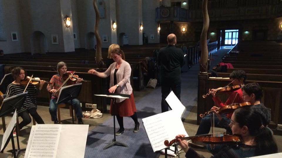 Ett antal fiolspelande personer sitter med notblad framför sig i en cirkel i kyrkan och en dirigent leder musiken. 