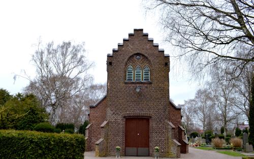 Råå kapell är byggt som en mindre kyrka i brunt tegel och grönt tak. I förgrunden syns buskar och träd.