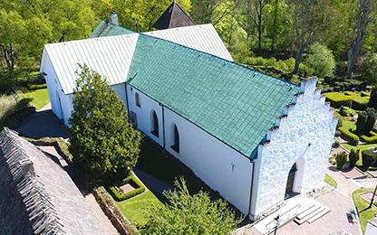 Ett flygfoto på en vit, medeltida kyrka. Kyrkan har grönt tak och trappstegsgavlar. Den ligger inbäddad i grönska.