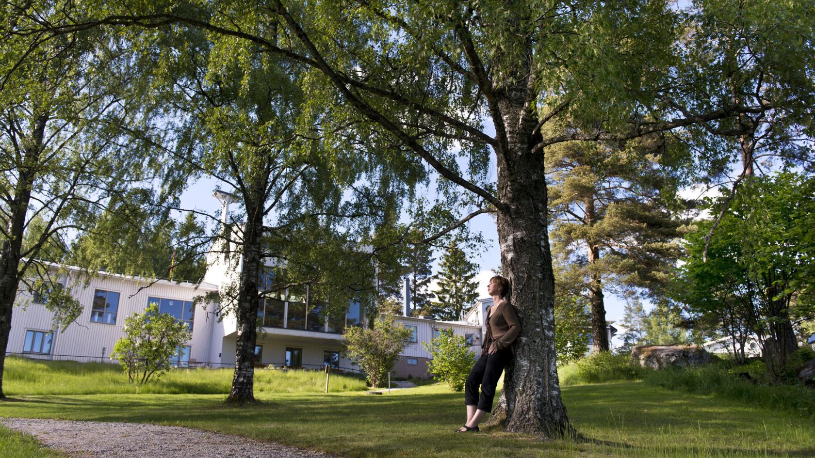 En kvinna lutar sig mot ett grönskande träd, i bakgrunden syns kursgården Solliden.